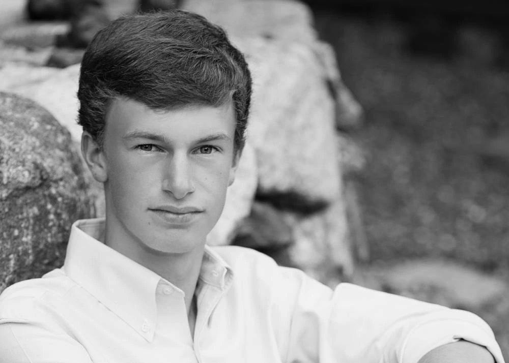 Black and white image of a young man sitting by a rock looking at the camera